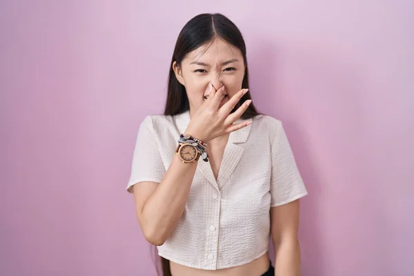中国の若い女性ピンクの背景の上に立って何か臭くて嫌な 耐え難い匂いを嗅ぎ 鼻の上に指で息を保持します 臭いが悪い — ストック写真