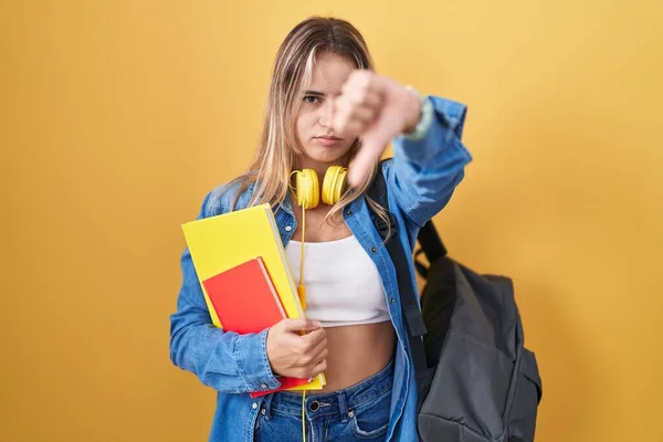 若いブロンドの女性は学生のバックパックを着て 親指を下にジェスチャーで拒絶と否定を示す不満と怒りを見て本を保持します 悪い表現 — ストック写真