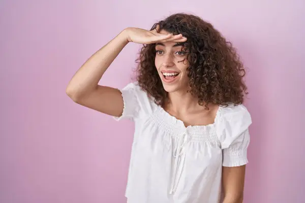 ピンクの背景の上に立っている巻き毛を持つヒスパニック系の女性は非常に幸せと笑顔遠くに手を頭の上に見て 検索の概念 — ストック写真