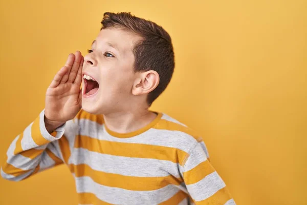 若い白人の子供は黄色の背景に立って大声で叫び 口の上に手で横に大声で叫んでいます コミュニケーションの概念 — ストック写真