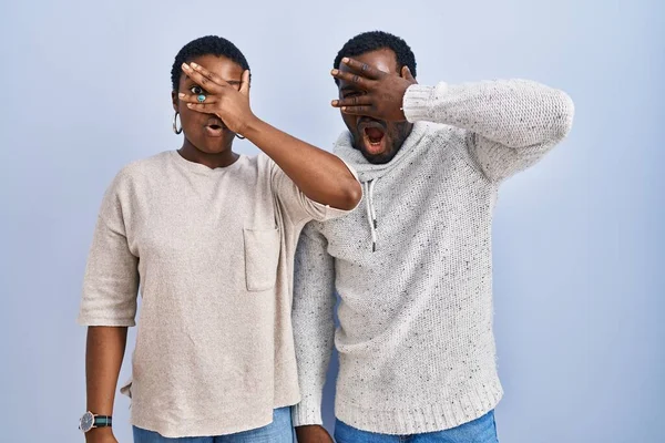 stock image Young african american couple standing over blue background together peeking in shock covering face and eyes with hand, looking through fingers with embarrassed expression. 