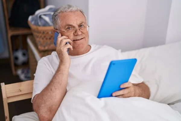 中世の白髪の男を使用してタッチパッドを使用してスマートフォン上で話してベッドルームに座って — ストック写真
