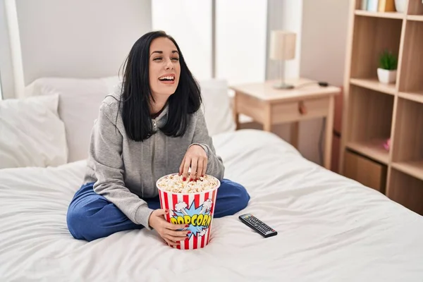 ヒスパニック系の女性食べるポップコーン見ますA映画上のベッド笑顔と笑いハードアウト大声でので面白いクレイジー冗談 — ストック写真