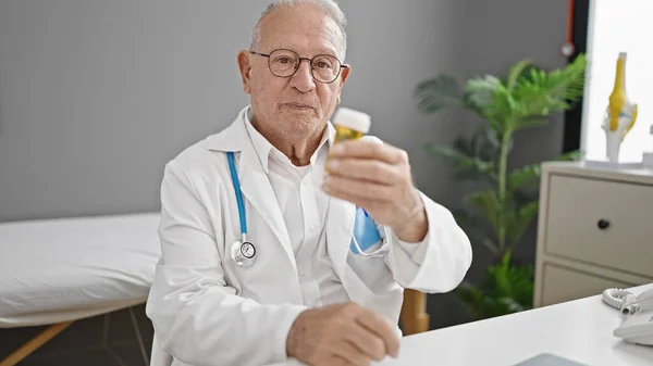 シニア白髪男医師保持丸薬上のテーブルに座ってクリニック — ストック写真