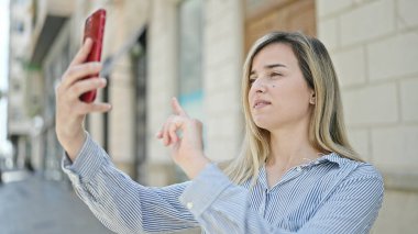 Genç sarışın kadın caddede akıllı telefonun yanında selfie çekiyor.