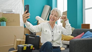 Orta yaşlı sarışın kadın yeni evindeki kanepede elinde anahtarla video görüşmesi yapıyor.