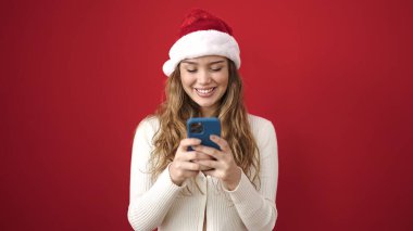 Genç, güzel, İspanyol bir kadın yılbaşı şapkası takıyor ve izole edilmiş kırmızı arka plan üzerinde akıllı telefon kullanıyor.