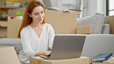 Genç kızıl saçlı kadın yeni evinde dizüstü bilgisayar kullanıyor.