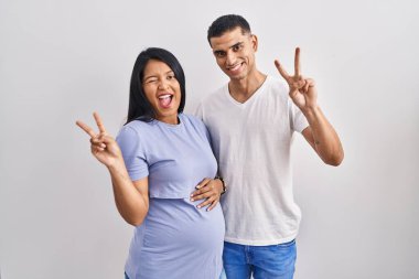 Latin kökenli genç bir çift arka planda durup kameraya gülümseyen ve parmaklarıyla zafer işareti yapan bir bebek bekliyor. İki numara.. 