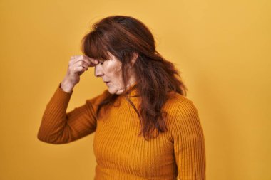 Orta yaşlı İspanyol kadın sarı arka planda duruyor. Burnu ve gözleri ovuşturmaktan yorulmuş, yorgunluk ve baş ağrısı çekiyor. Stres ve hüsran kavramı. 