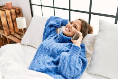 Genç, güzel, İspanyol bir kadın yatakta akıllı telefondan konuşuyor.