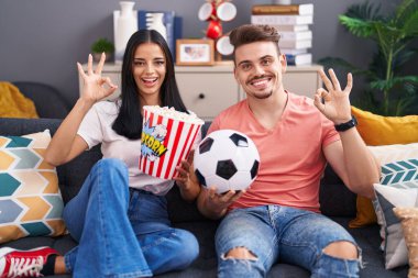 Genç İspanyol çift futbol holiganları top tutuyorlar ve patlamış mısır yiyorlar parmaklarıyla işaret veriyorlar, gülümseyerek mükemmel bir sembol oluşturuyorlar. 