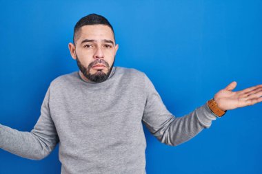 Hispanic man standing over blue background clueless and confused expression with arms and hands raised. doubt concept. 