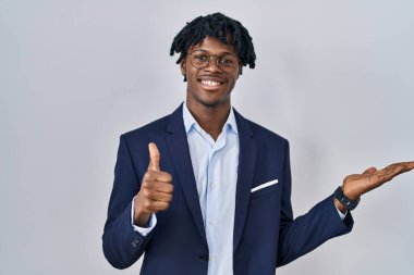 Beyaz arka planda iş ceketi giyen Afrikalı genç bir adam avuç içini gösteriyor ve baş parmaklarıyla hareket ediyor, mutlu ve neşeli gülümsüyor. 