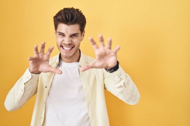 Young hispanic man standing over yellow background smiling funny doing claw gesture as cat, aggressive and sexy expression 