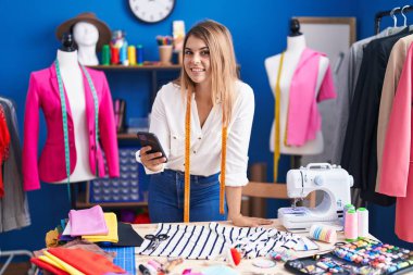 Young woman tailor smiling confident using smartphone at sewing studio