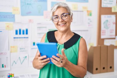 Orta yaşlı, gri saçlı, iş kadını. Ofiste dokunmatik ped kullanarak gülümsüyor.