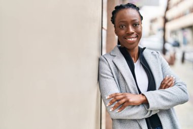 Afro-Amerikalı kadın gülümsüyor. Kollarını kavuşturup caddede duruyor.