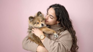 Köpeğiyle gülümseyen genç İspanyol kadın pembe arka planda duruyor.