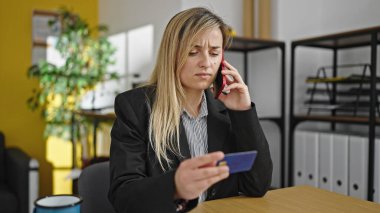 Young blonde woman business worker talking on smartphone holding credit card at office