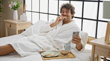 Genç İspanyol adam kahvaltısını yapıyor. Yatak odasında akıllı telefondan video izliyor.