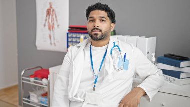 Afro-Amerikalı erkek doktor, klinikte rahat bir şekilde masada oturuyor.