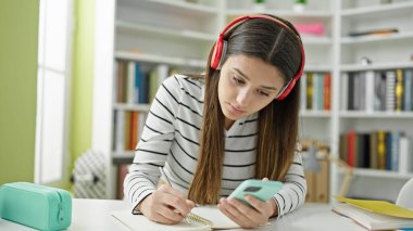 Genç ve güzel İspanyol öğrenci kütüphane üniversitesinde akıllı telefon ve kulaklık kullanarak notlar yazıyor.