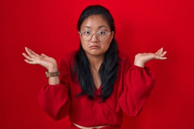 Asyalı genç bir kadın kırmızı arka planda duruyor. Kolları ve elleri havada, kafası karışmış bir ifade. şüphe kavramı. 