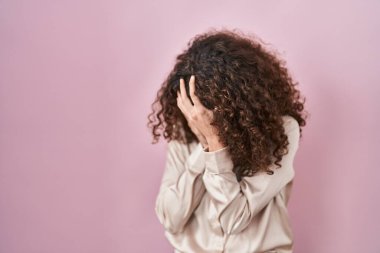 Hispanic woman with curly hair standing over pink background with sad expression covering face with hands while crying. depression concept. 