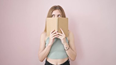 Genç sarışın kadın, pembe arka plan üzerine kitap ile ağzını kapatıyor.