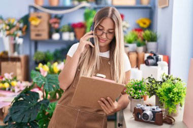 Young beautiful hispanic woman florist talking on smartphone reading clipboard at flower shop