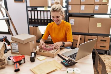 Genç sarışın kadın ecommerce işçisi ofisindeki karton kutuda diyadem paketliyor.