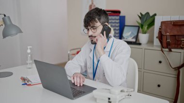 Genç İspanyol doktor bilgisayarını kullanarak klinikte telefonla konuşuyor.