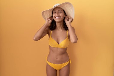 Bikini giyen, yaz şapkalı, gülümseyen kulakları parmaklarıyla çeken İspanyol bir kadın. Komik bir jest. deneme sorunu 