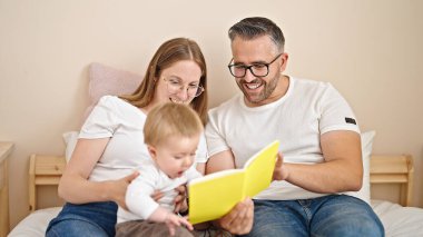 Anne, baba ve bebeğin ailesi yatak odasındaki yatakta oturmuş kitap okuyor.