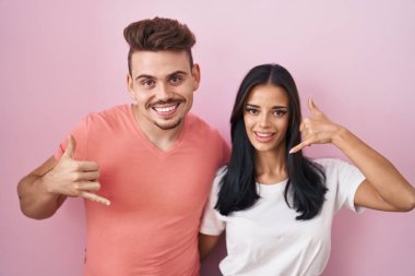Pembe arka planda durup gülümseyen genç İspanyol çift telefonla el ve parmaklarıyla telefonla konuşuyor gibi davranıyorlar. iletişim kavramları. 