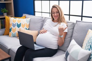 Evde kanepede oturan genç hamile bir kadın dizüstü bilgisayarını kullanarak başparmağıyla yan tarafa bakıyor ve ağzı açık gülümsüyor. 