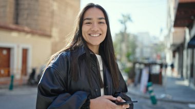 Genç, güzel, İspanyol bir kadın sokakta akıllı telefon kullanarak gülümsüyor.