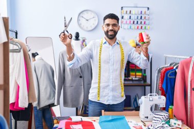 Young arab man tailor smiling confident holding scissors and thread at tailor shop