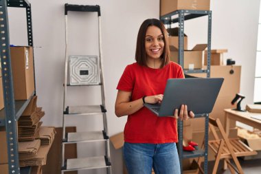 Young beautiful hispanic woman ecommerce business worker using laptop standing at office