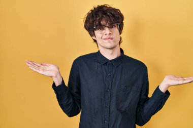 Sarı arka planda gözlük takan genç adam elleri ve kolları havada, kafası karışık bir ifade sergiliyor. şüphe kavramı. 