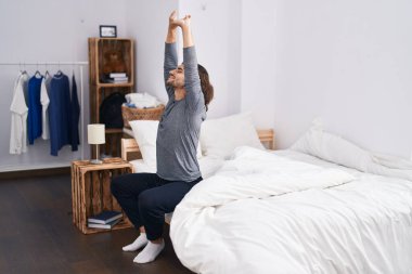Young hispanic man waking up stretching arms at bedroom