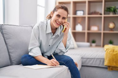 Not defterine yazan genç bir kadın evde akıllı telefondan konuşuyor.