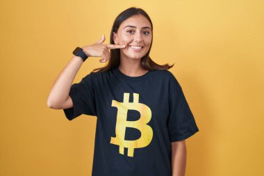 Bitcoin tişörtü giyen, gülümseyen, parmaklarıyla ve ağzıyla işaret eden genç İspanyol kadın. diş sağlığı kavramı. 