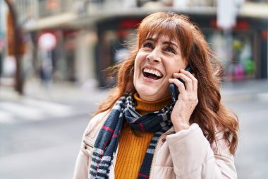 Orta yaşlı bir kadın gülümsüyor. Sokakta akıllı telefondan konuşuyor.