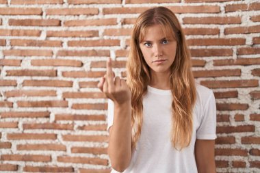 Young caucasian woman standing over bricks wall showing middle finger, impolite and rude fuck off expression 