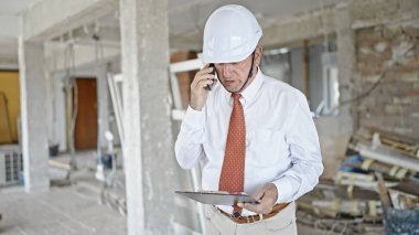 Kıdemli gri saçlı mimar inşaat alanında akıllı telefon okuma raporuyla konuşuyor.