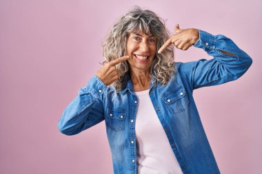 Middle age woman standing over pink background smiling cheerful showing and pointing with fingers teeth and mouth. dental health concept. 