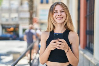 Sokakta gülümseyen akıllı telefon kullanan genç sarışın kadın.