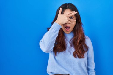 Hispanic young woman standing over blue background peeking in shock covering face and eyes with hand, looking through fingers with embarrassed expression. 
